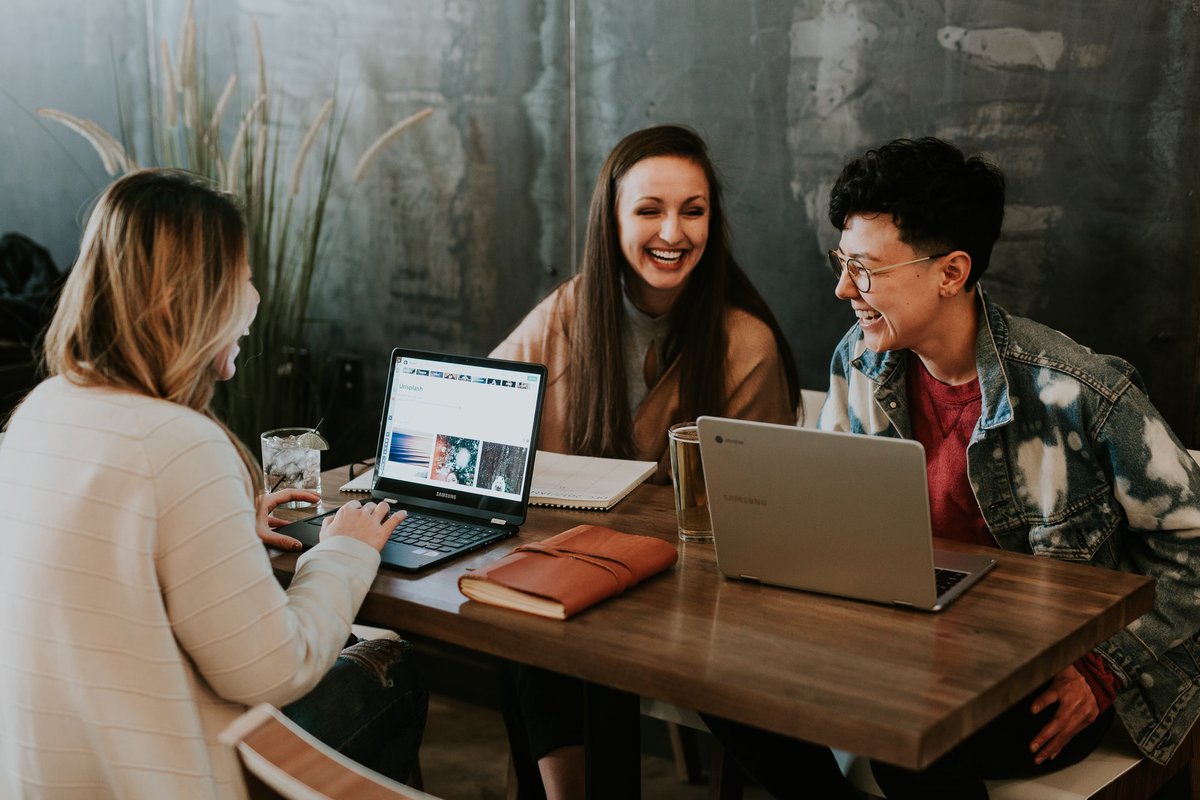 Three people laugh as they work at laptops on a shared bench